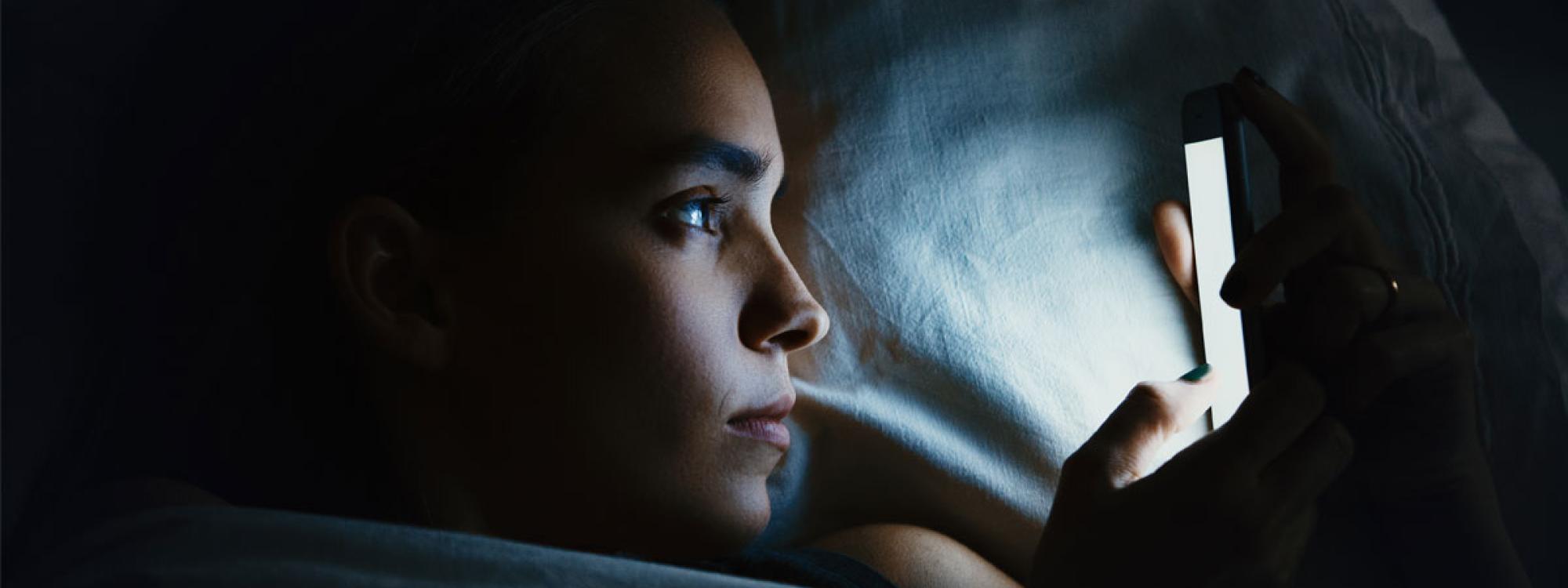 Woman laying in bed in darkness, looking at illuminated phone screen